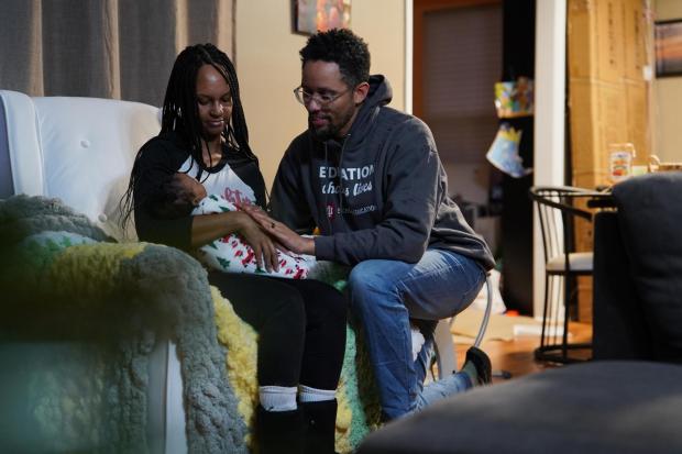 Crystal Wilmot-Burton holds their sleeping newborn daughter as her husband, JaKobi Burton, kneels next to her.
