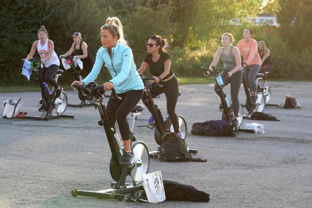 A woman pedals on a stationary exercise bike with others during a spinning class in a parking lot 