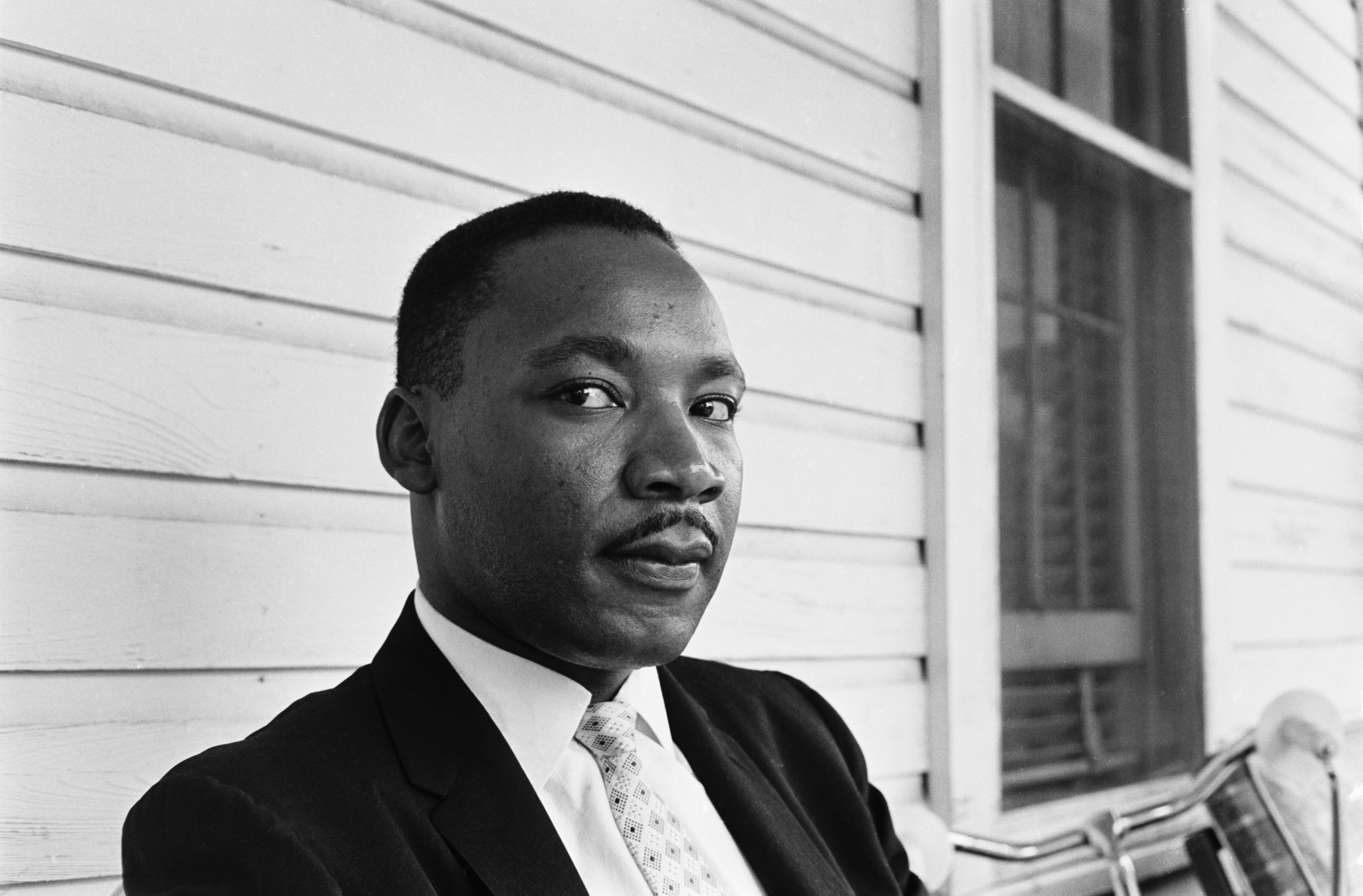 In this black and white portrait of The Rev. Dr. Martin Luther King, Jr., he wears a suit and tie and looks pensively at the camera. 