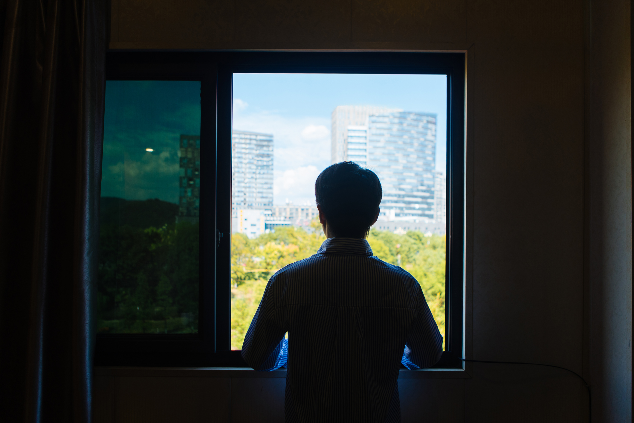 Young man in China standing in front of a window looking into the distance, with a green park and modern skyscrapers outside.