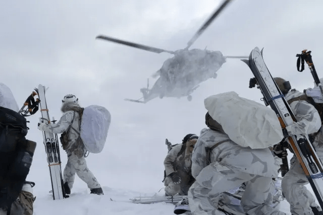 Soldiers in snowy camouflage carrying skis approach a helicopter in a blizzard.