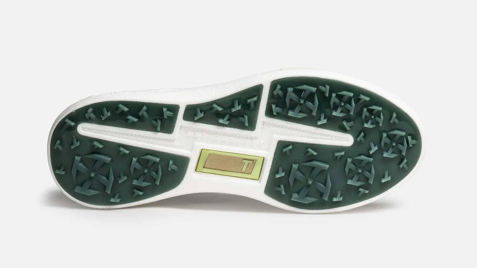 The image shows the outsole of a True Linkswear golf shoe, featuring a white base with green rubber tread sections. The tread has geometric patterns and arrows, and there is a small rectangular gold plate in the center with a T on it.