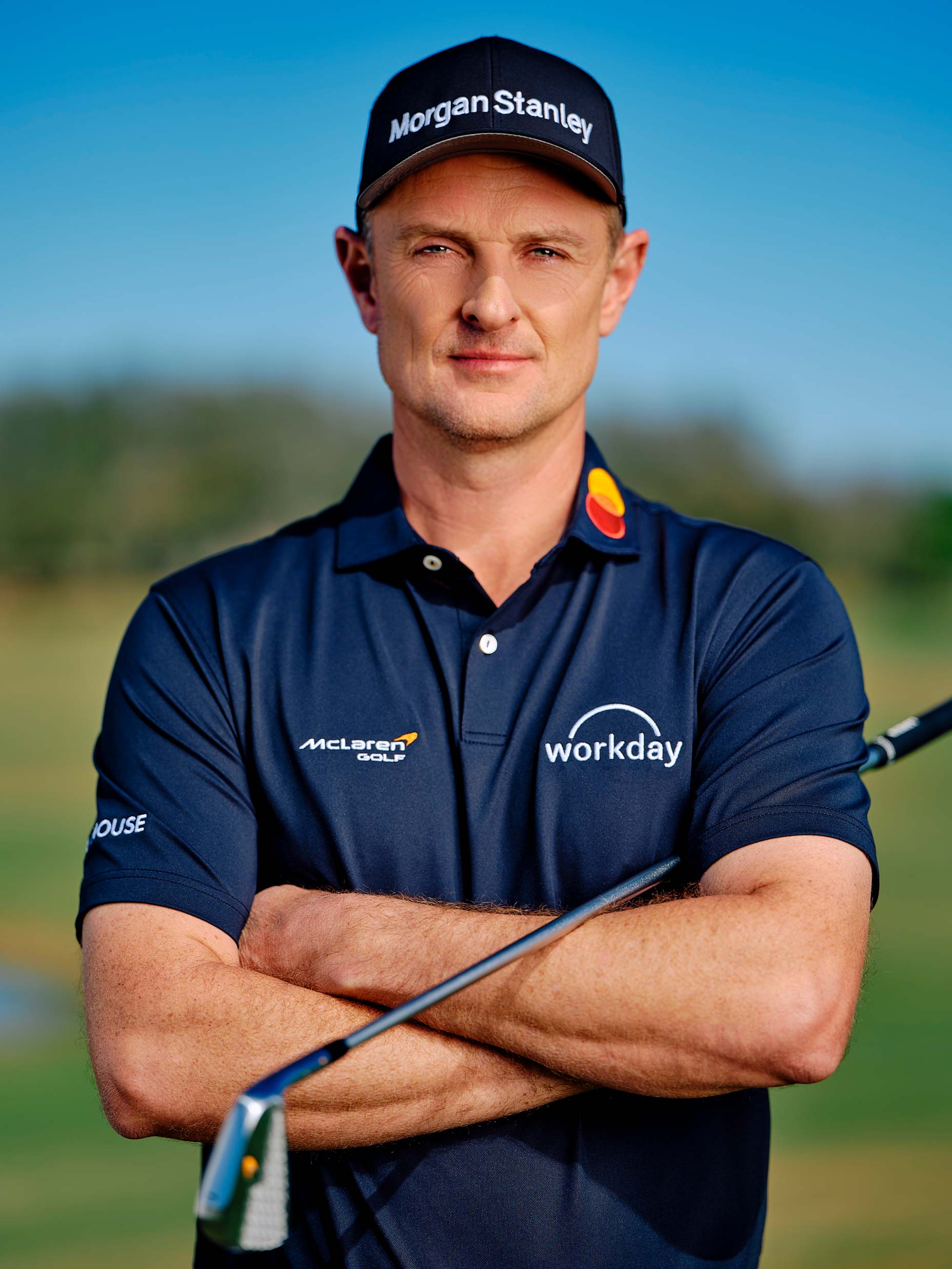 A male golfer, Justin Rose, stands outdoors with arms crossed, wearing a navy blue polo shirt and a cap branded with Morgan Stanley and Workday. He holds a golf club, with a golf course and blue sky in the background&mdash;a scene after his shocking move to McLaren.