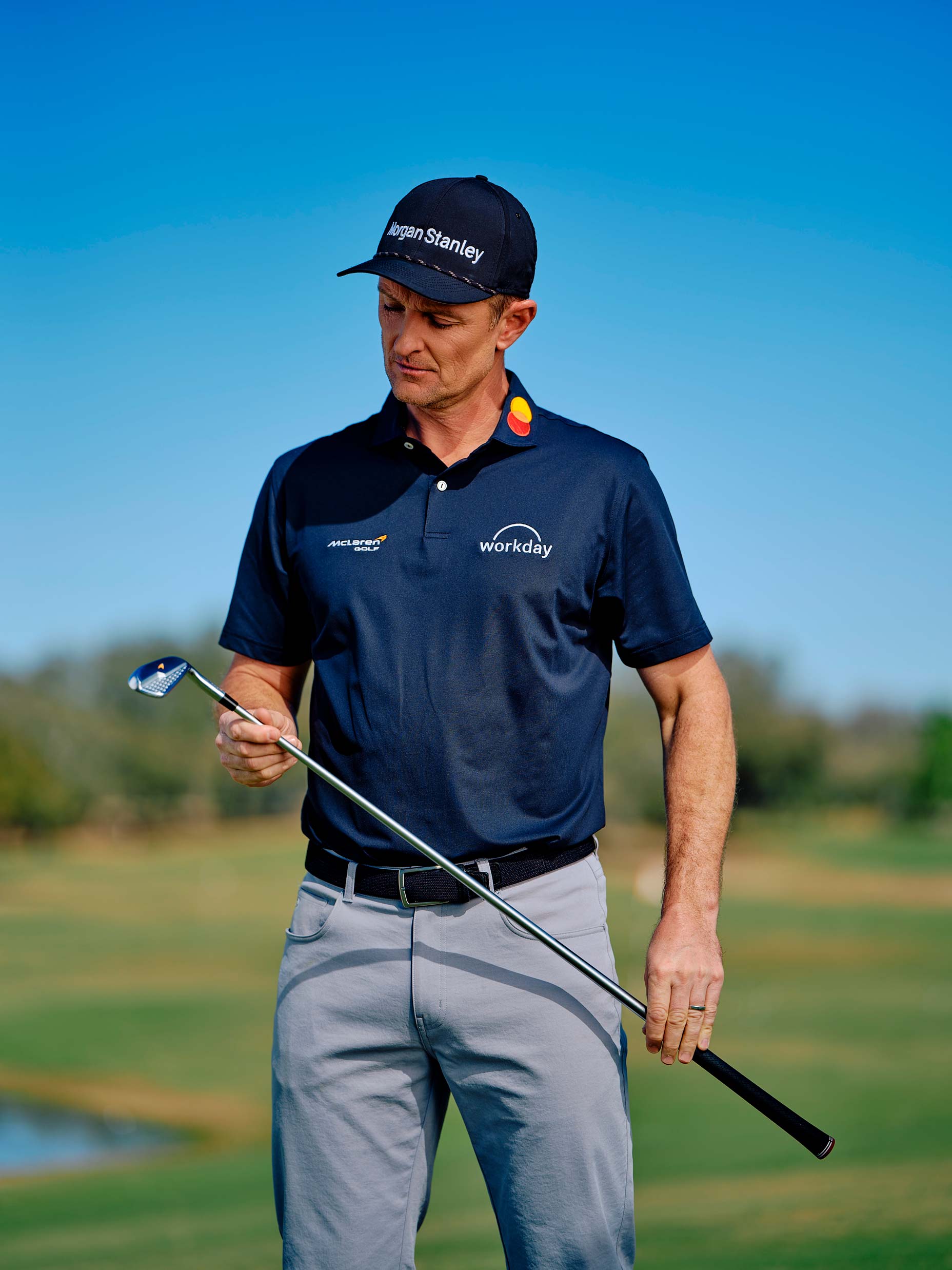 A golfer wearing a navy cap and polo with sponsor logos, including McLaren, stands on a golf course, looking down thoughtfully at a golf club held in his hands. The background shows green grass and blue sky.
